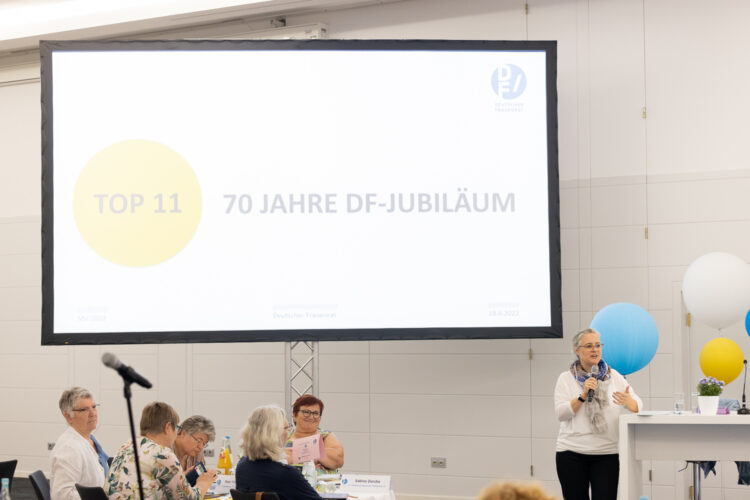 Bildschirm mit "70 Jahre Deutscher Frauenrat", rechts davon Anja Nordmann mit Mikro, im Vordergrund Delegierta am Tisch